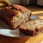A loaf of moist sugar free banana bread with one slice cut and resting beside it on a white plate.