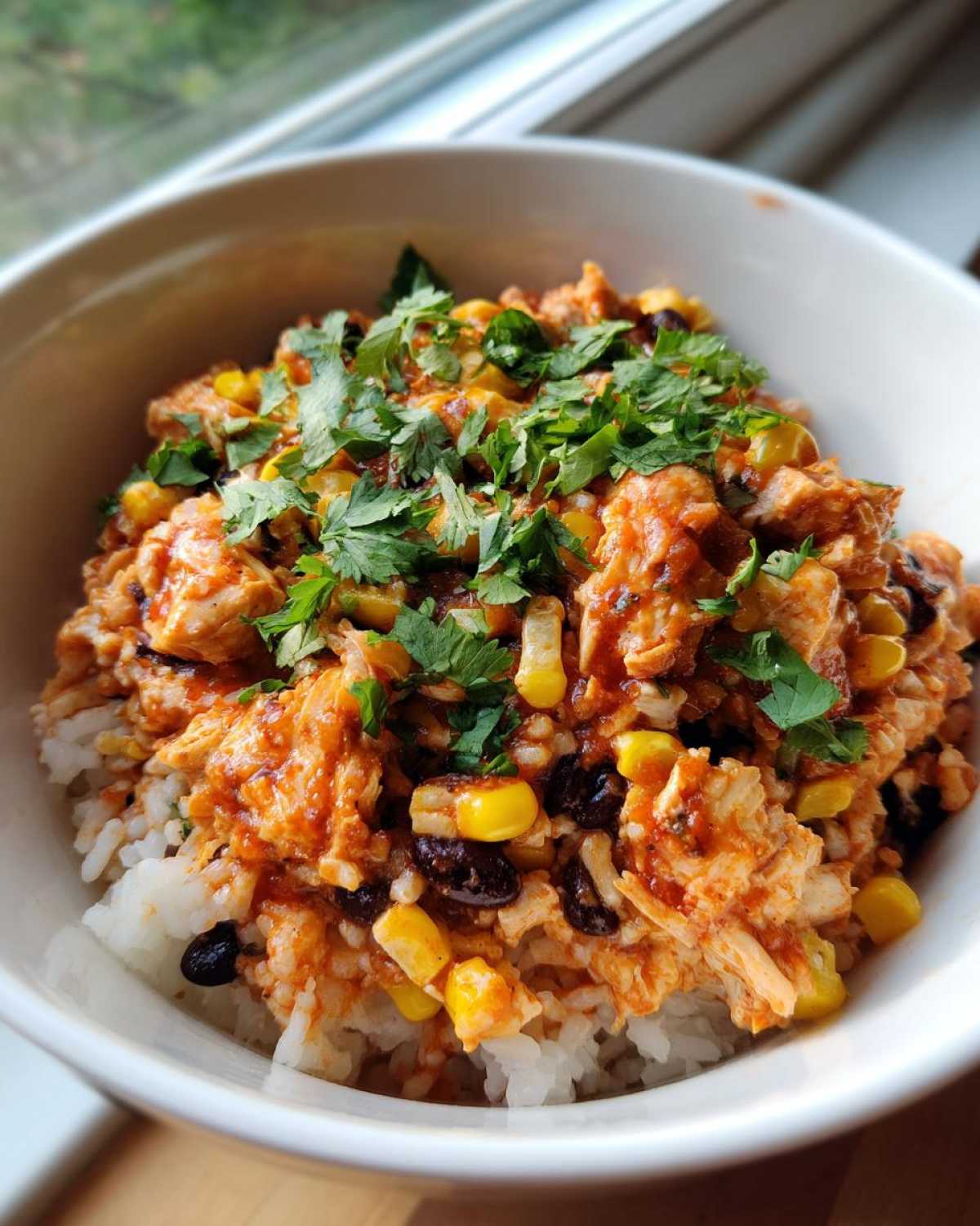A close-up shot of a white bowl filled with a delicious chicken burrito bowl, featuring rice, saucy chicken, corn, black beans, and fresh cilantro.