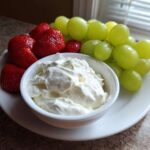 A bowl of creamy white fruit dip served on a white plate alongside fresh strawberries and green grapes.
