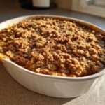 Close-up of a freshly baked apple crisp recipe with a golden, crunchy topping in a white oval baking dish.