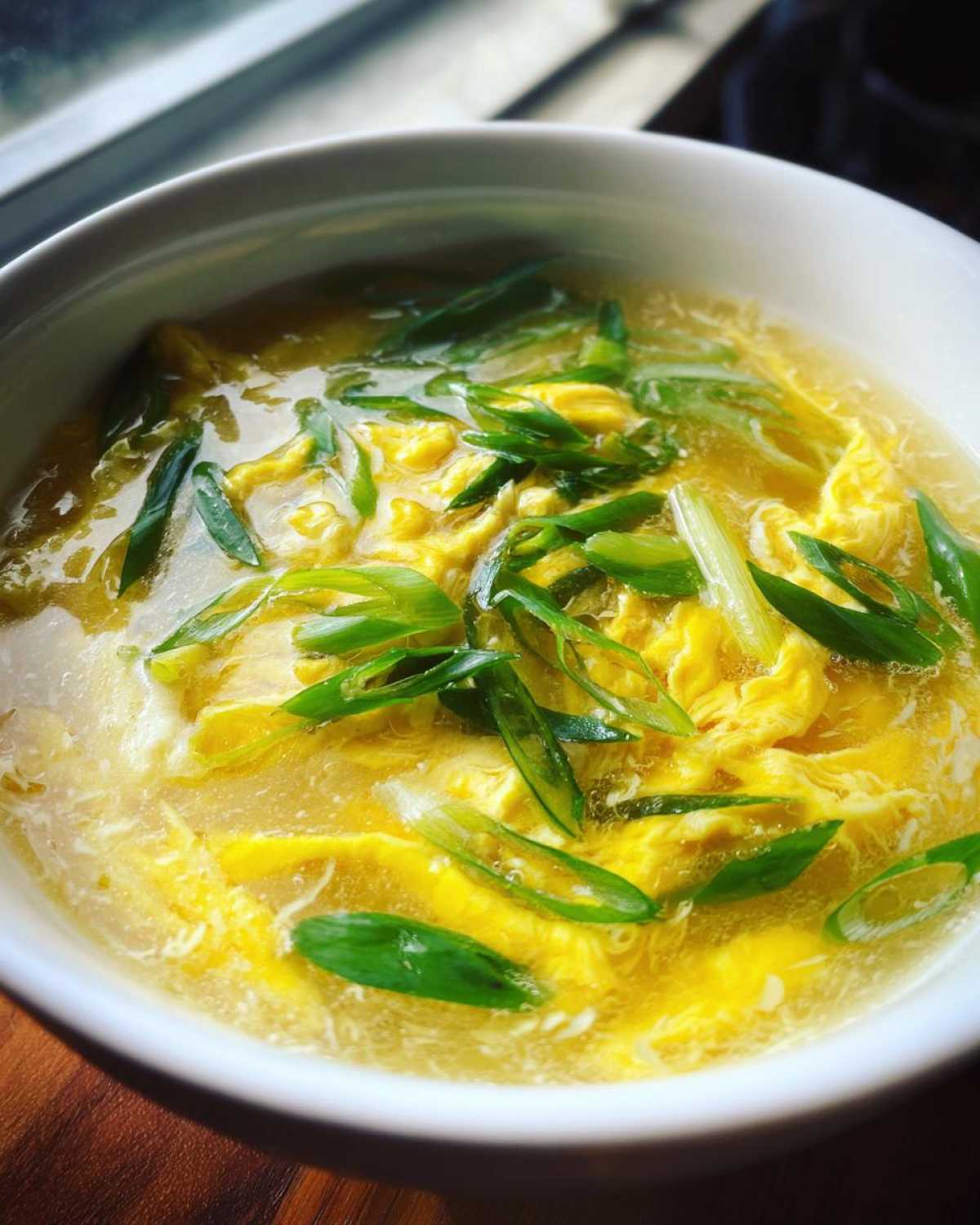 A close-up of a white bowl filled with bright yellow egg drop soup, garnished generously with fresh green scallions.
