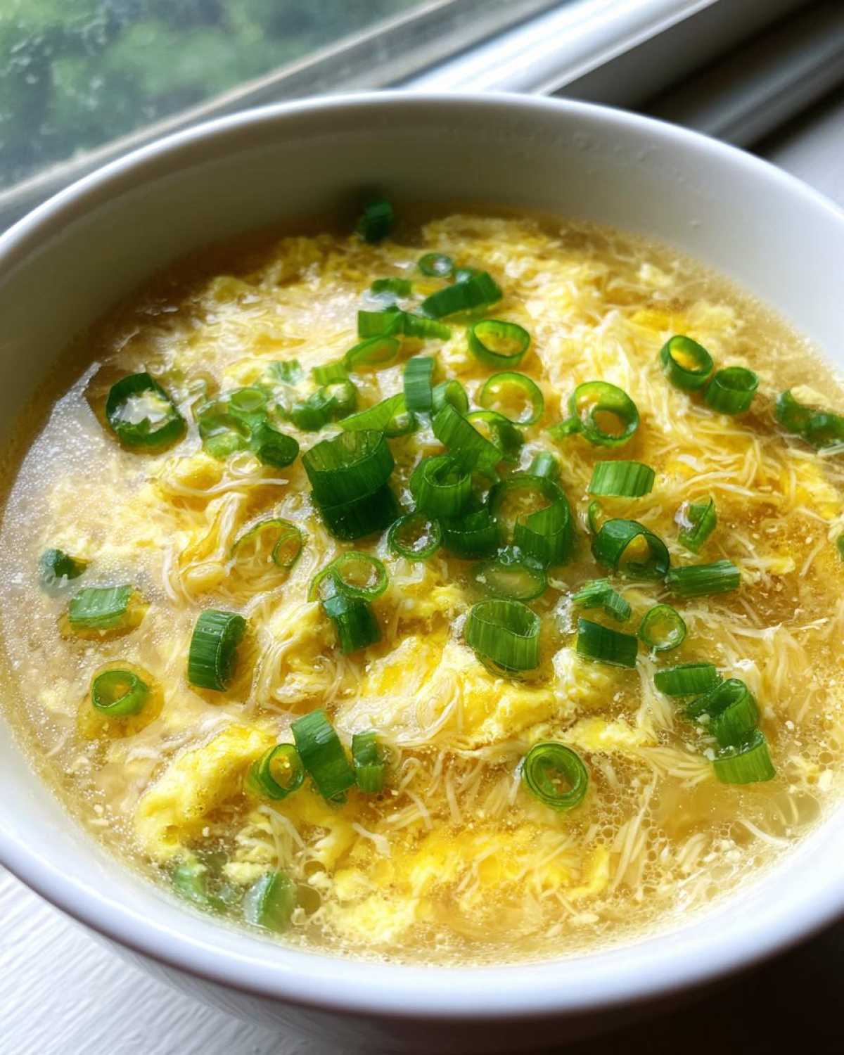 A close-up view of hot egg drop soup with delicate ribbons of cooked egg and topped generously with bright green sliced scallions.