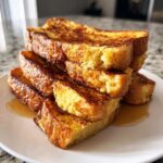 A tall stack of thick-cut, golden brown french toast drizzled with syrup on a white plate.