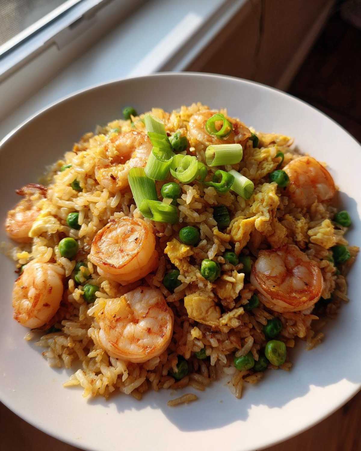 A bright, close-up shot of homemade shrimp fried rice with visible shrimp, peas, and egg, garnished with scallions.