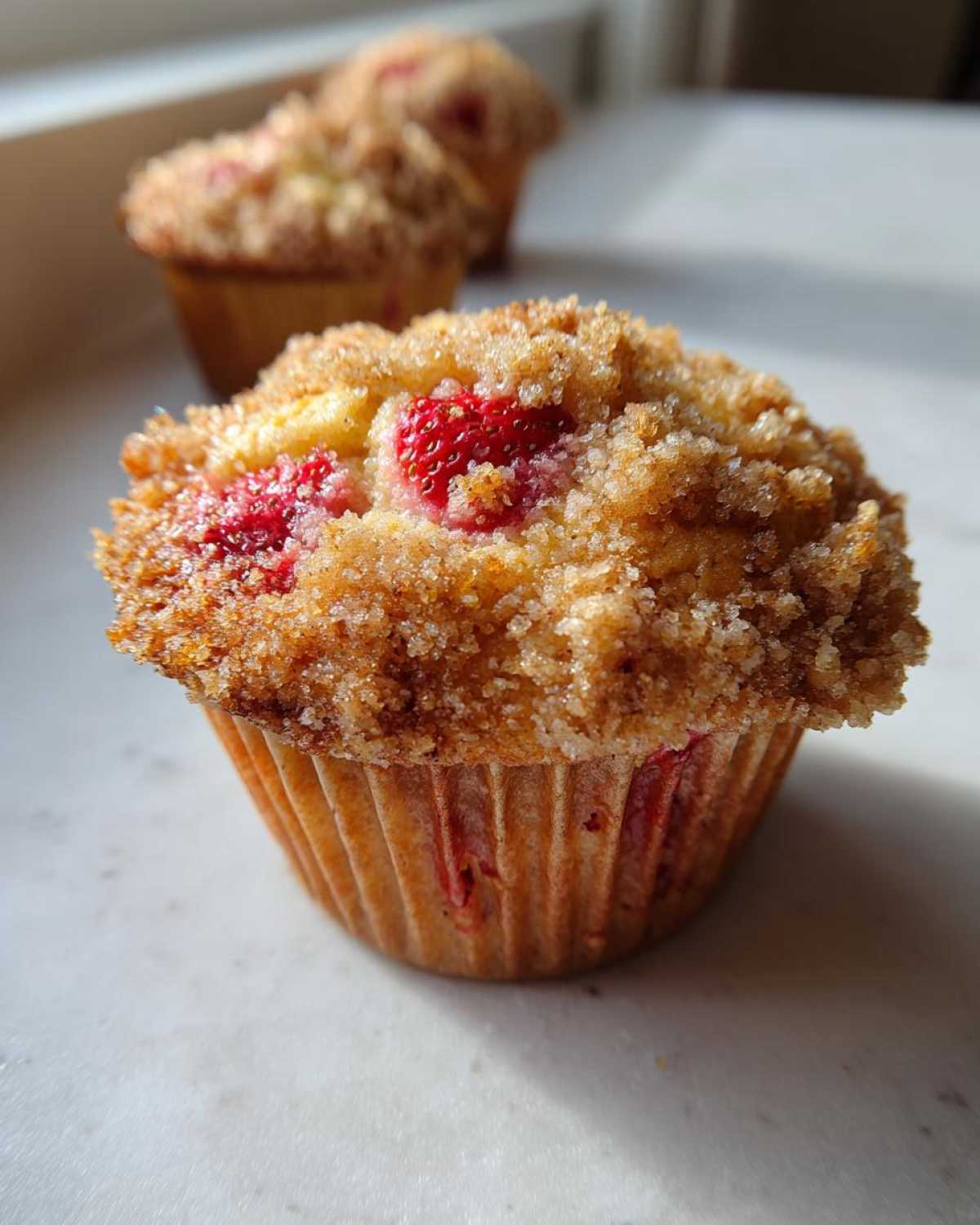 A close-up of a freshly baked strawberry muffin topped with a thick, sugary crumb topping and visible pieces of strawberry.