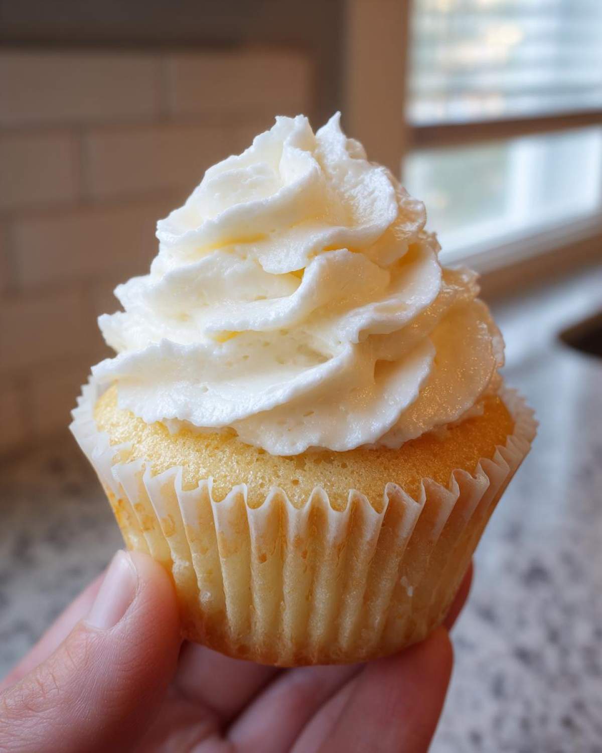 Close-up of a perfectly baked vanilla cupcakes topped with high swirl of white frosting, held by a hand.
