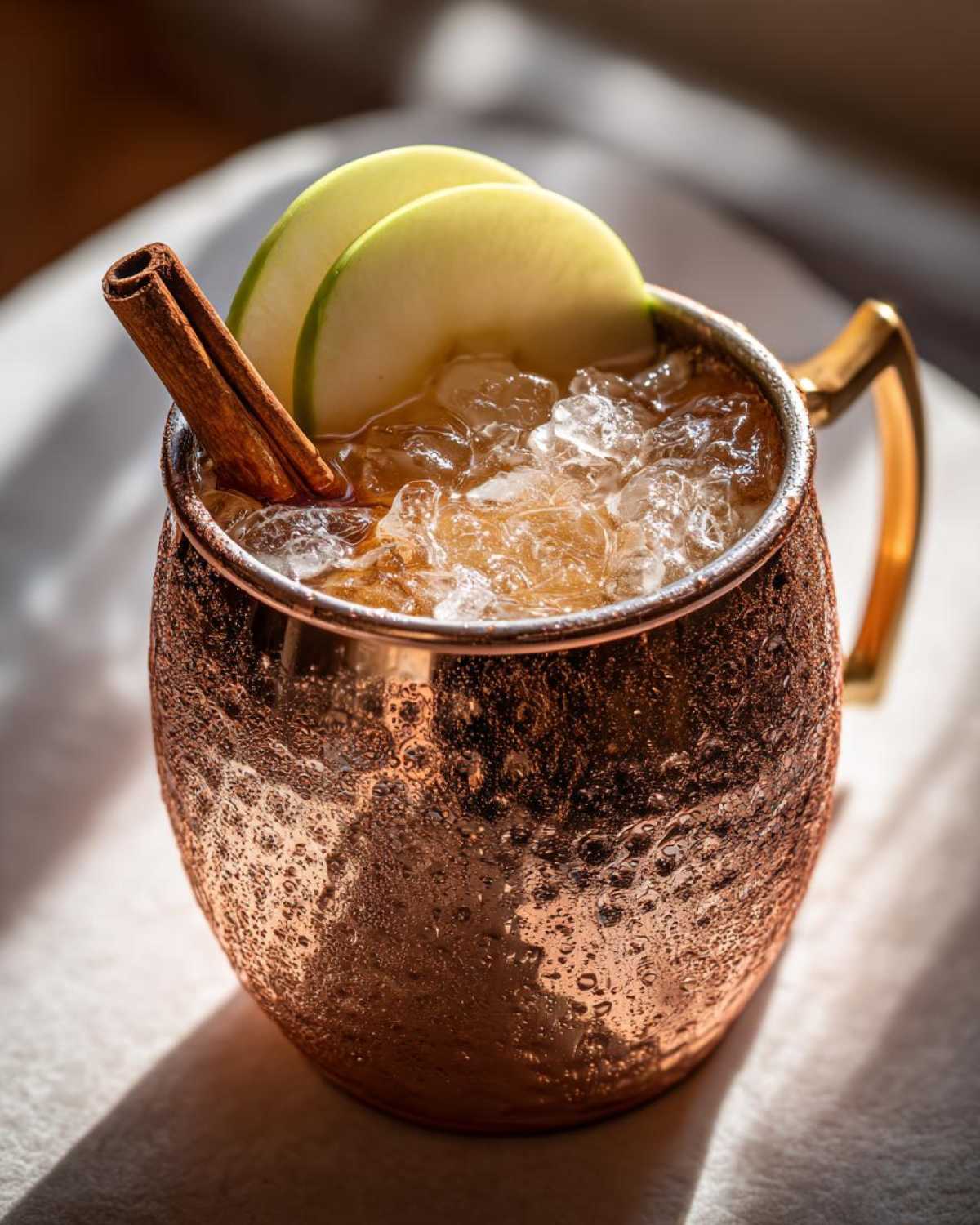 Close-up of a frosty apple cider moscow mule in a copper mug, garnished with apple slices and a cinnamon stick.
