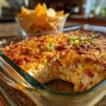 Close-up of a baked Seven layer dip in a glass dish with a scoop removed, topped with melted cheese and green onions.