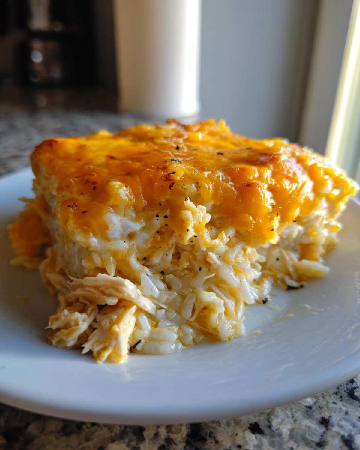 A square serving of cheesy chicken and rice bake topped with melted cheddar cheese on a white plate.