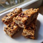 Close-up of four chewy no bake granola bars stacked on a white plate, featuring oats, chocolate chips, and dried fruit.