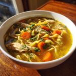 Close-up of a white bowl filled with rich chicken noodle soup, featuring egg noodles, shredded chicken, carrots, and parsley.