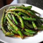 A close-up of bright green, glossy chinese green beans with garlic sauce piled on a white plate.