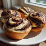 Four golden-brown chocolate swirl pinwheel cookies arranged on a white plate, catching bright sunlight.