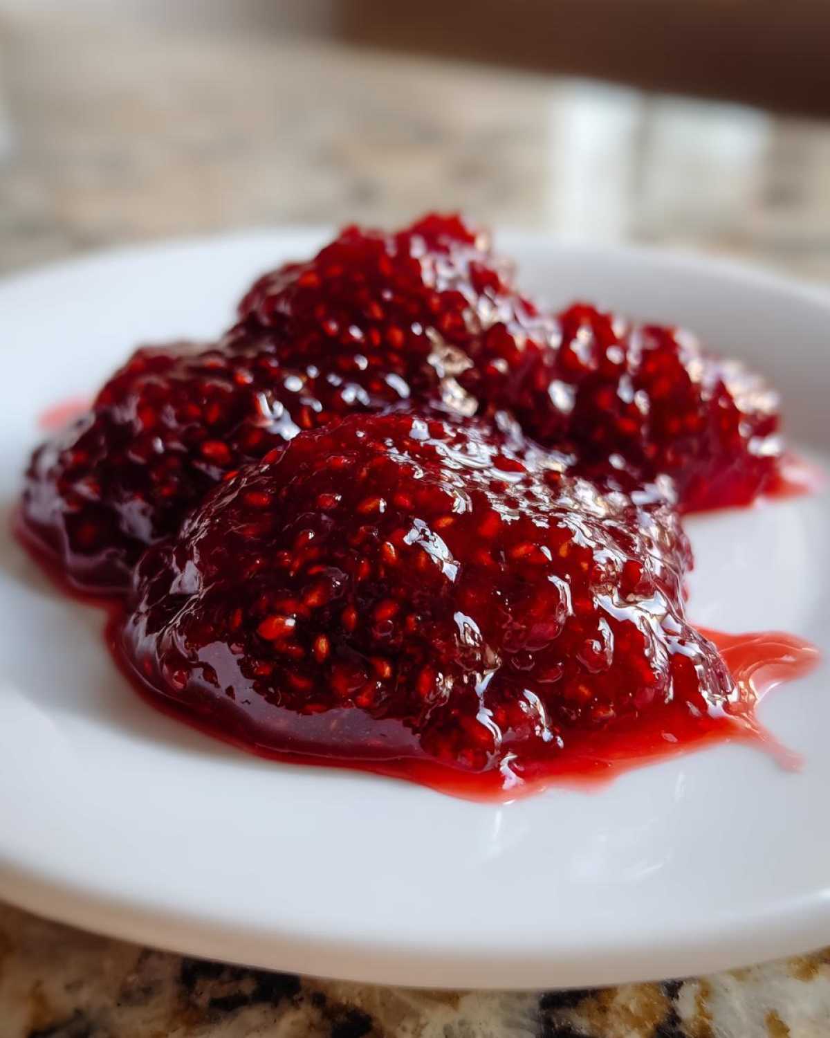 Three dollops of vibrant, chunky raspberry jam with visible seeds resting on a white plate.