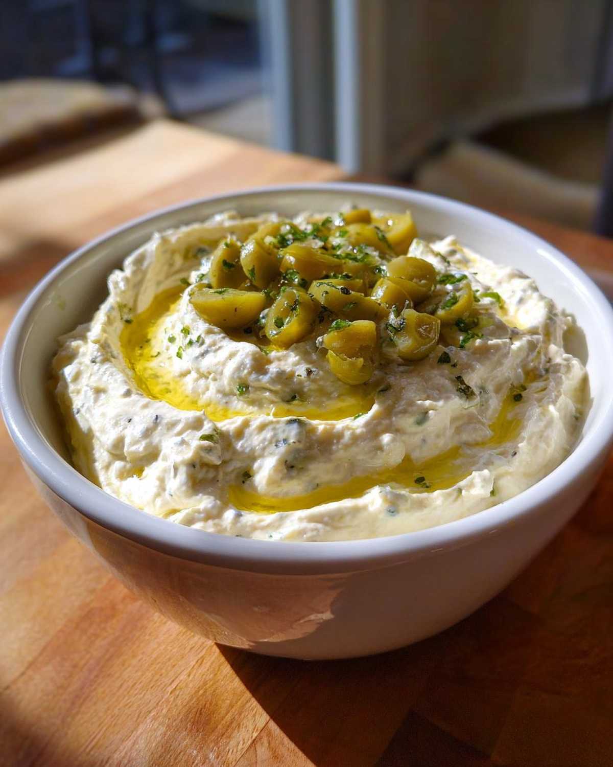 A bowl of creamy dirty martini dip garnished with sliced green olives and olive oil.