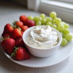 A bowl of creamy white fruit dip surrounded by fresh strawberries and green grapes on a white plate.