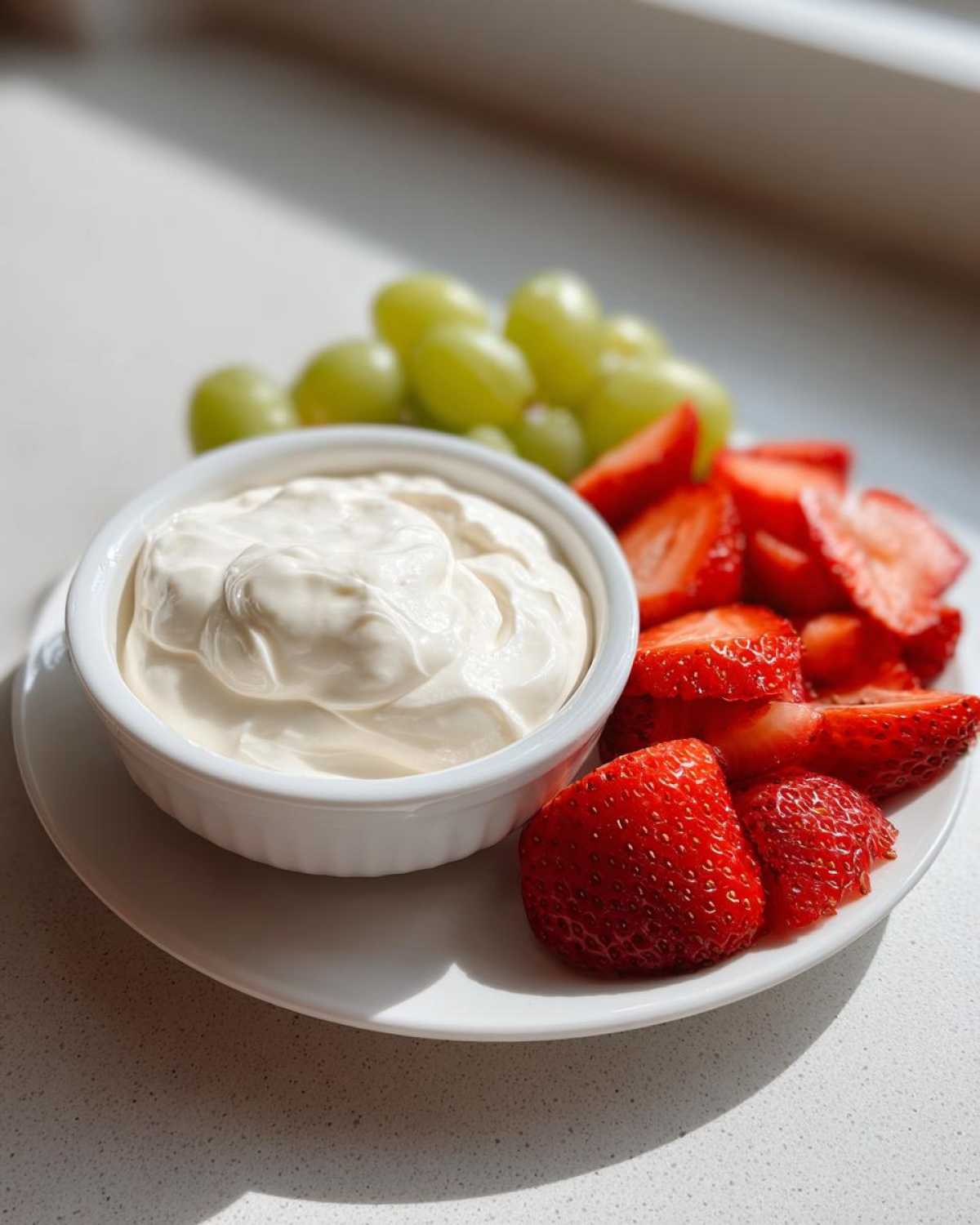 A small white bowl of creamy fruit dip served with sliced strawberries and green grapes.