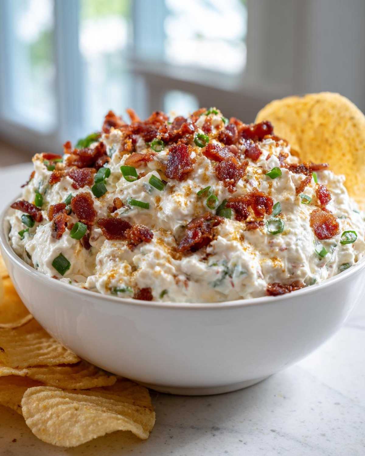 A white bowl filled with creamy Million Dollar Dip, topped generously with crumbled bacon and chopped green onions.