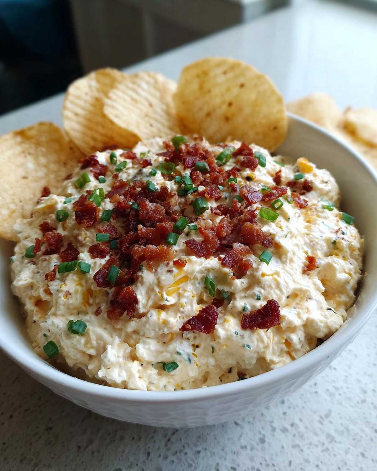 A bowl of creamy Million Dollar Dip topped generously with crispy bacon bits and fresh green chives, served with ridged potato chips.