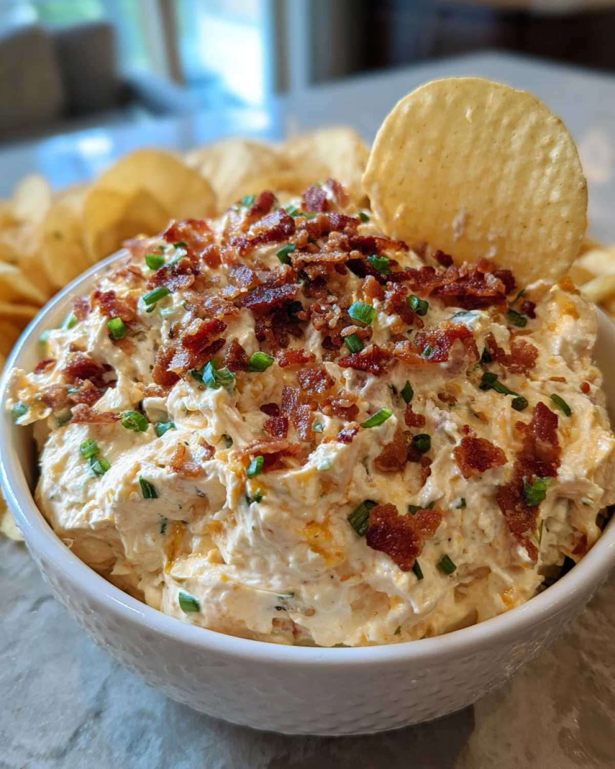 A bowl of creamy Million Dollar Dip topped generously with crumbled bacon and fresh chives, served with a potato chip.