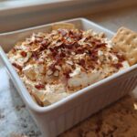 A close-up of creamy Neiman Marcus Dip topped with crumbled bacon and sliced almonds, served with crackers.