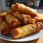 A stack of freshly fried, golden brown, crispy egg roll appetizers piled high on a white plate.