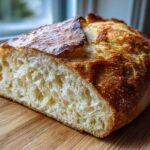 A loaf of crusty Italian bread, sliced open to show the airy, open crumb structure, resting on a wooden cutting board.