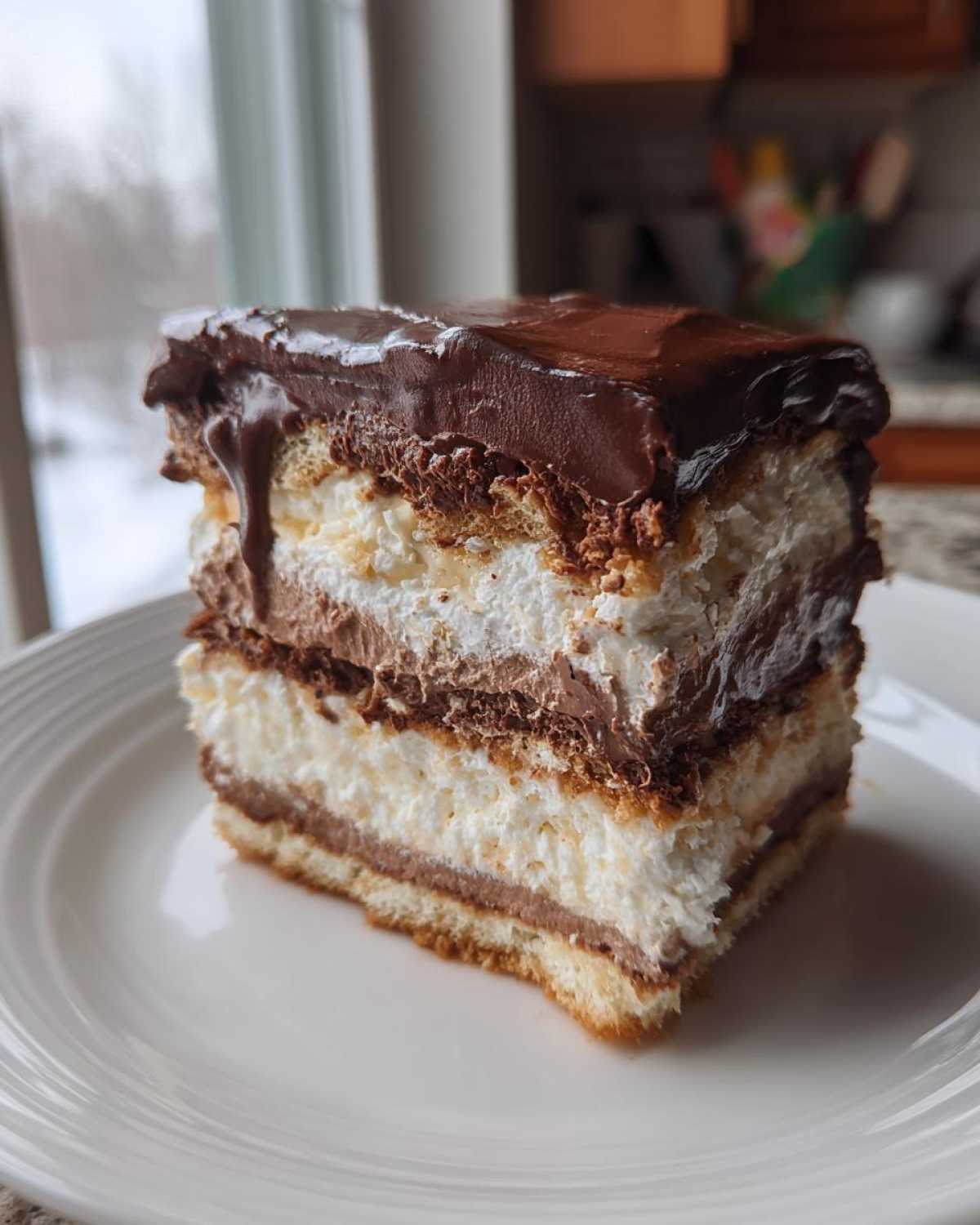 A tall, layered slice of eclair cake with vanilla cream, chocolate pudding, and a thick chocolate glaze on top.