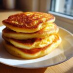 A stack of four golden brown, fluffy coconut flour pancakes served on a white plate near a window.