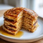 A tall stack of fluffy oatmeal pancakes with a piece cut out, drizzled generously with maple syrup.