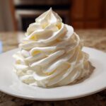 A tall swirl of fluffy, pale yellow vanilla buttercream frosting piped onto a small white plate.