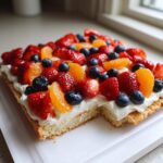 A rectangular fruit pizza sugar cookie topped with white frosting, strawberries, blueberries, and orange slices, with one piece removed.