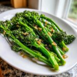A close-up of bright green, perfectly cooked broccolini tossed with browned garlic pieces, served on a white platter.