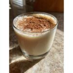 A close-up of a glass filled with creamy coquito, generously dusted with ground cinnamon on top.