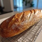 A freshly baked, golden brown french bread loaf with crisp scoring resting on a wire cooling rack.