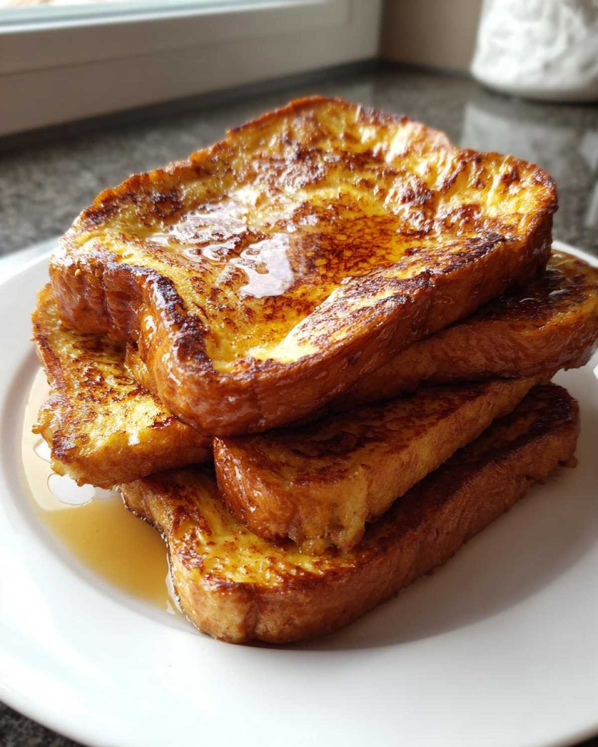 A stack of four golden brown slices of french toast drizzled generously with maple syrup on a white plate.