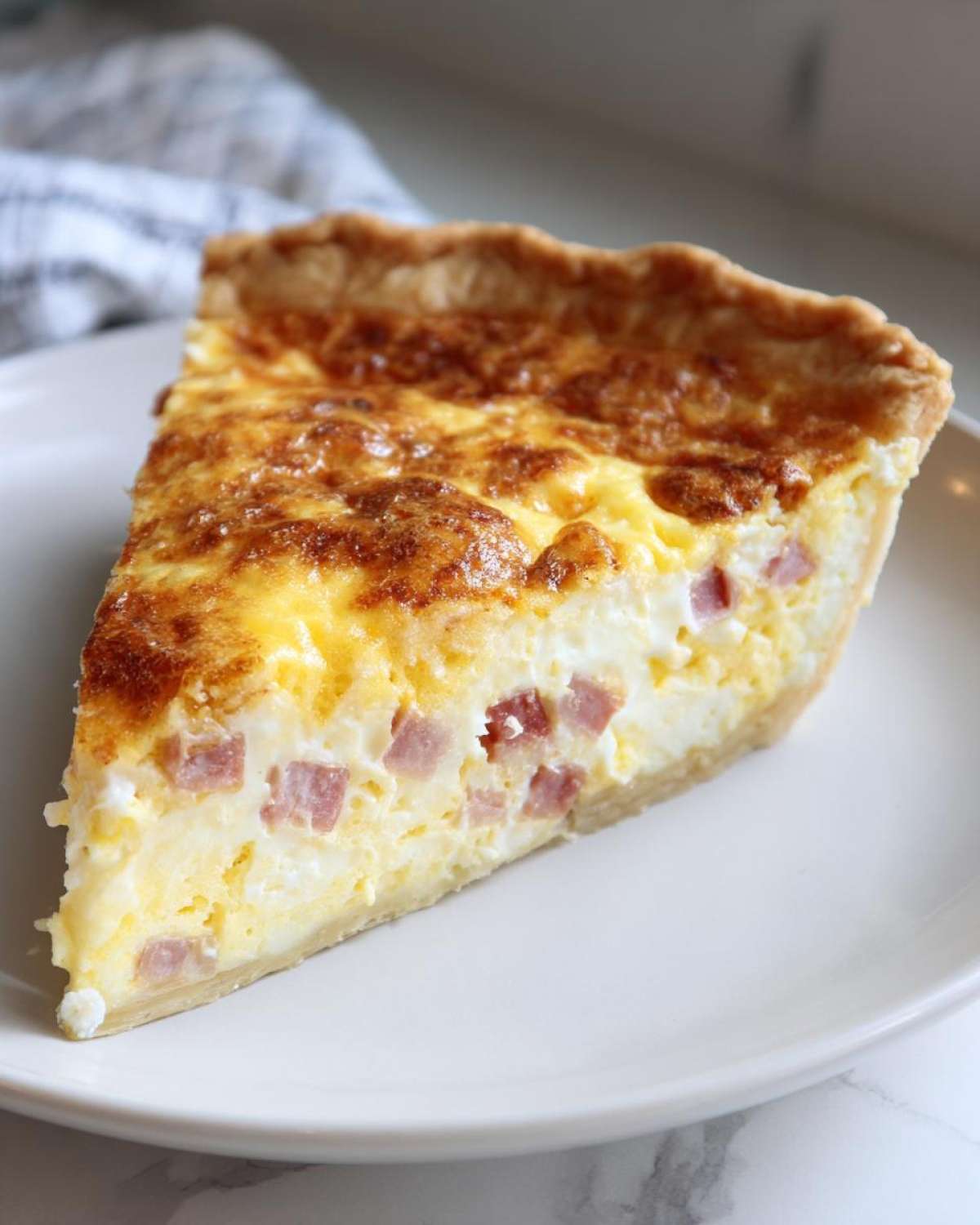 A perfect slice of ham and cheese quiche on a white plate, showing the rich egg filling and cubed ham.