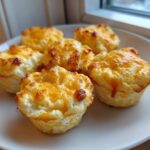 Five fluffy, baked egg bites topped with melted, golden-brown cheese served on a white plate.