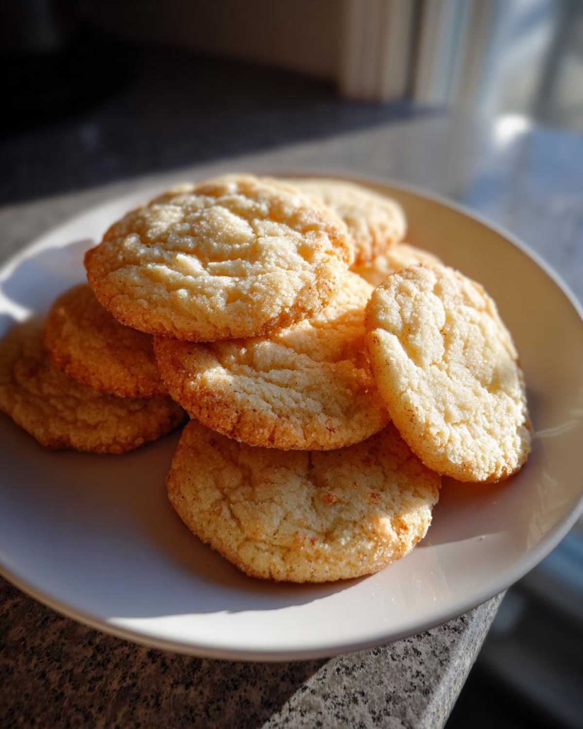 A stack of freshly baked, golden brown butter cookie treats resting on a light-colored plate near a window.