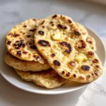 A stack of warm homemade naan bread, topped with melting pats of butter, showing charred spots.