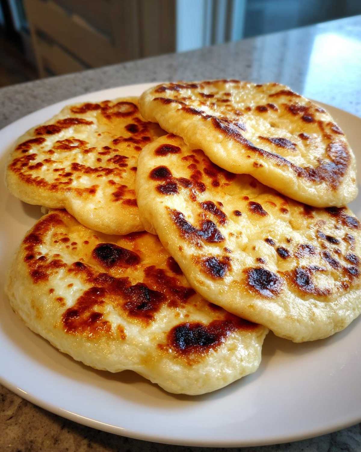 A stack of four freshly cooked homemade naan bread rounds with golden-brown, slightly charred spots, served on a white plate.