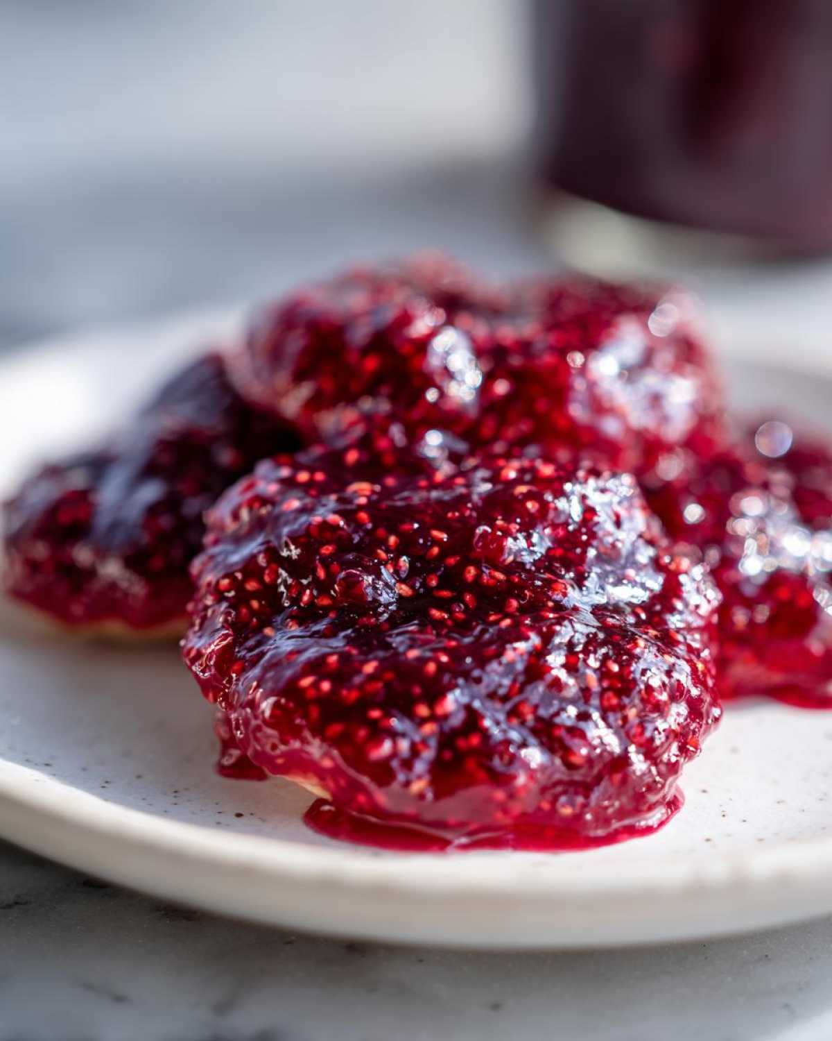 Close-up of thick, glossy, deep red raspberry jam with visible seeds spread on small biscuits or scones.