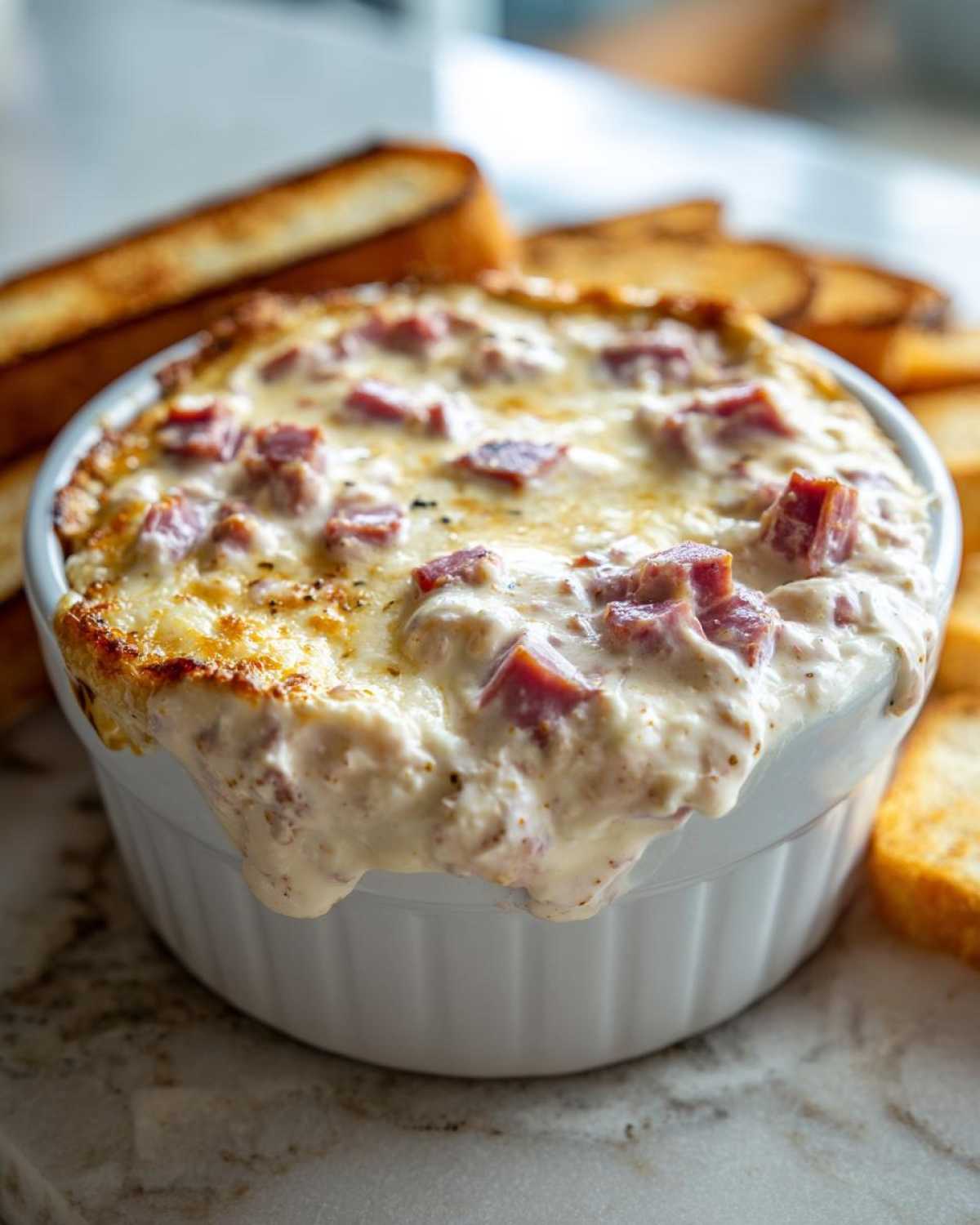 Close-up of a hot, bubbly reuben dip overflowing slightly from a white ramekin, served with toasted bread.