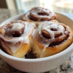 Three gooey, freshly baked cinnamon rolls topped with white icing served in a small white bowl.