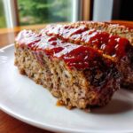 Close-up of thick slices of juicy meatloaf topped with a shiny, thick red glaze, served on a white plate.