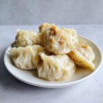A close-up of several juicy Traditional Chinese Dumplings piled on a small white plate against a light background.