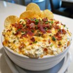 A close-up of hot, cheesy Million Dollar Dip topped with crispy bacon bits and fresh green chives, served with tortilla chips.
