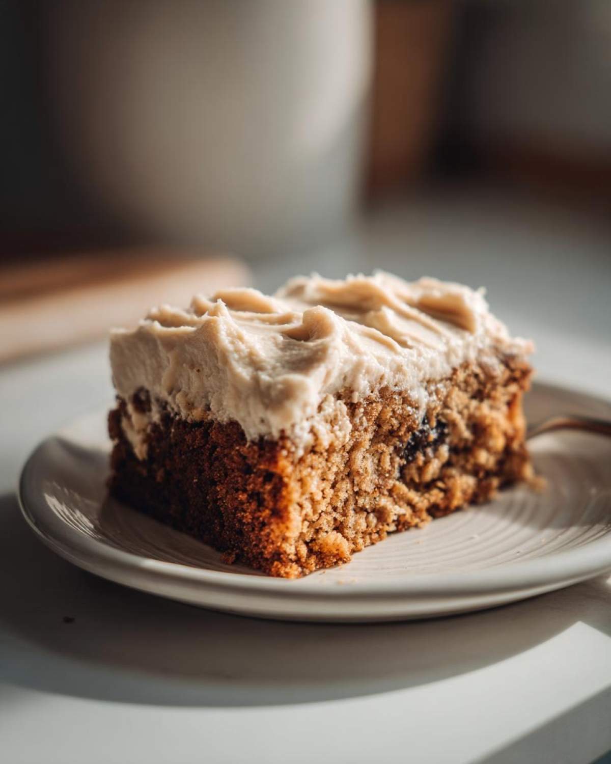 Close-up of a moist slice of applesauce cake topped with thick, swirled frosting on a small white plate.