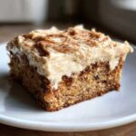 A close-up of a moist slice of applesauce cake topped with thick, creamy frosting and a dusting of cinnamon.