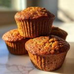 A close-up stack of three moist bran muffins with golden-brown tops, beautifully lit by sunlight.